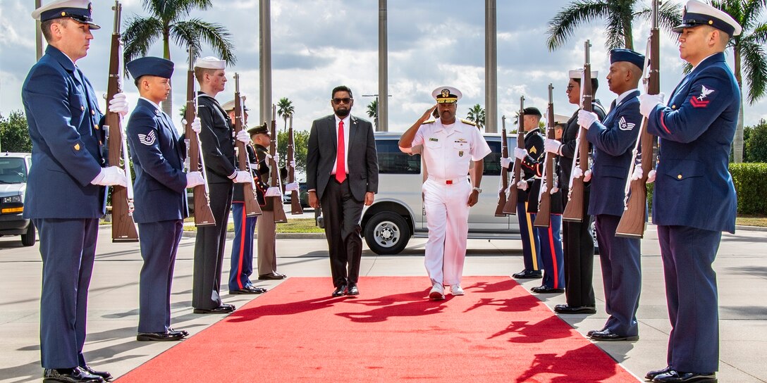 A man in a white military uniform walks on a red carpet aside a man in a blue suit and flanked on each side by a military color guard.