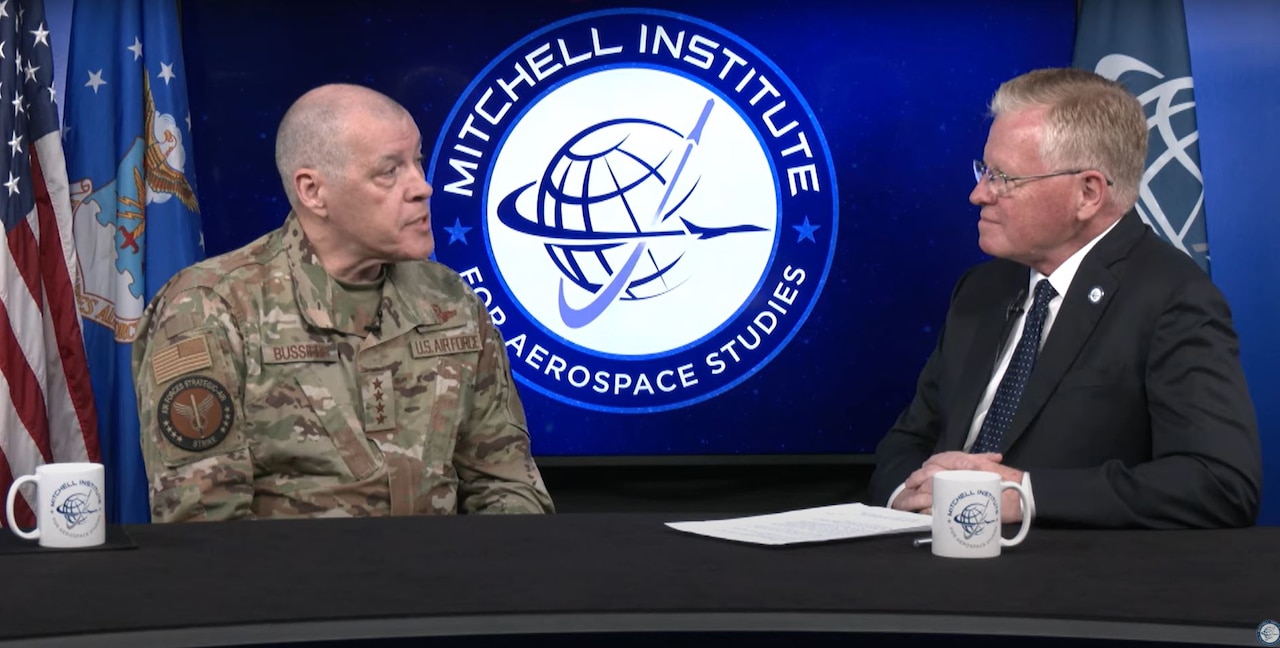 Two men, one in military camouflage and the other in a business suit, are sitting at a desk having a conversation.