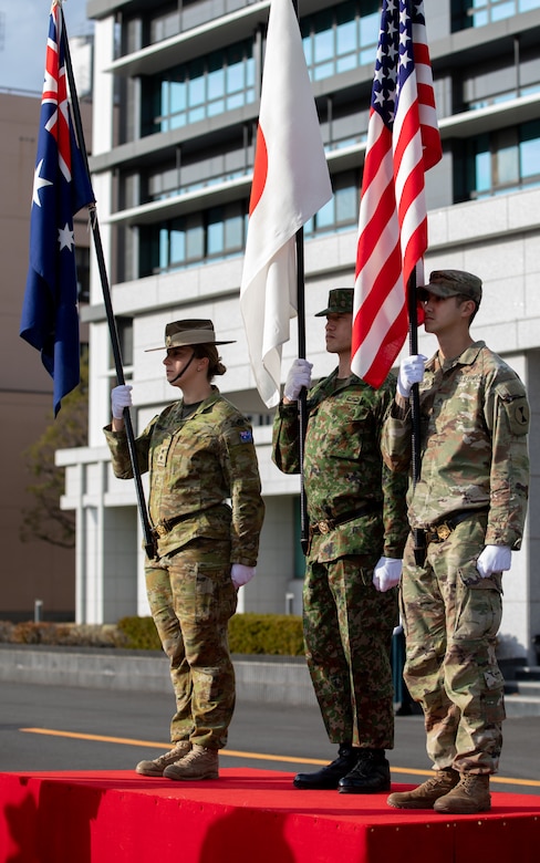 Three service members hold flags of the United States, Japan and Australia in front of a building.