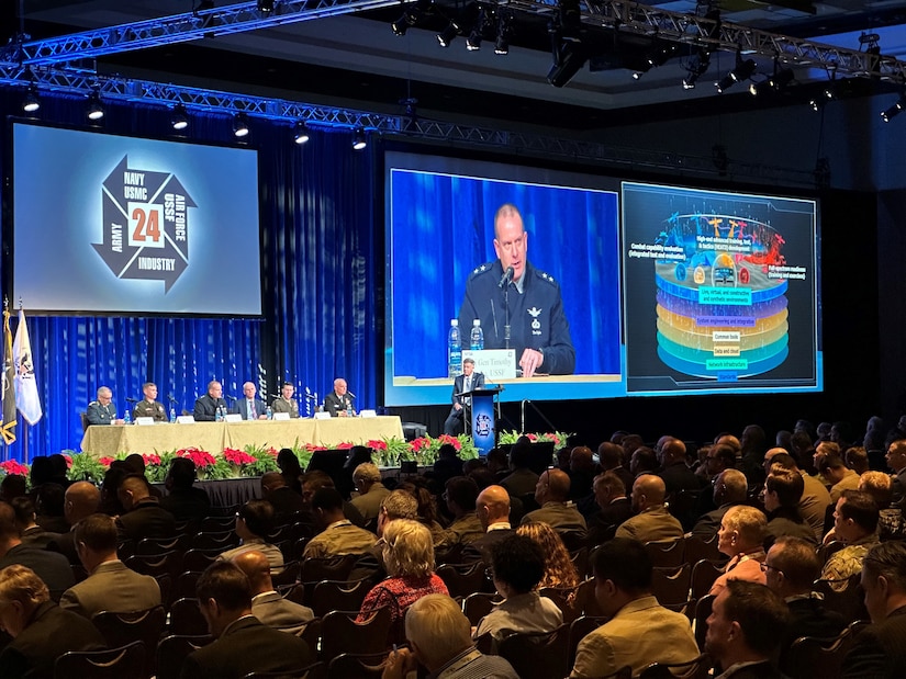 A large screen shows a view of a guardian speaking in a large room with a seated audience.