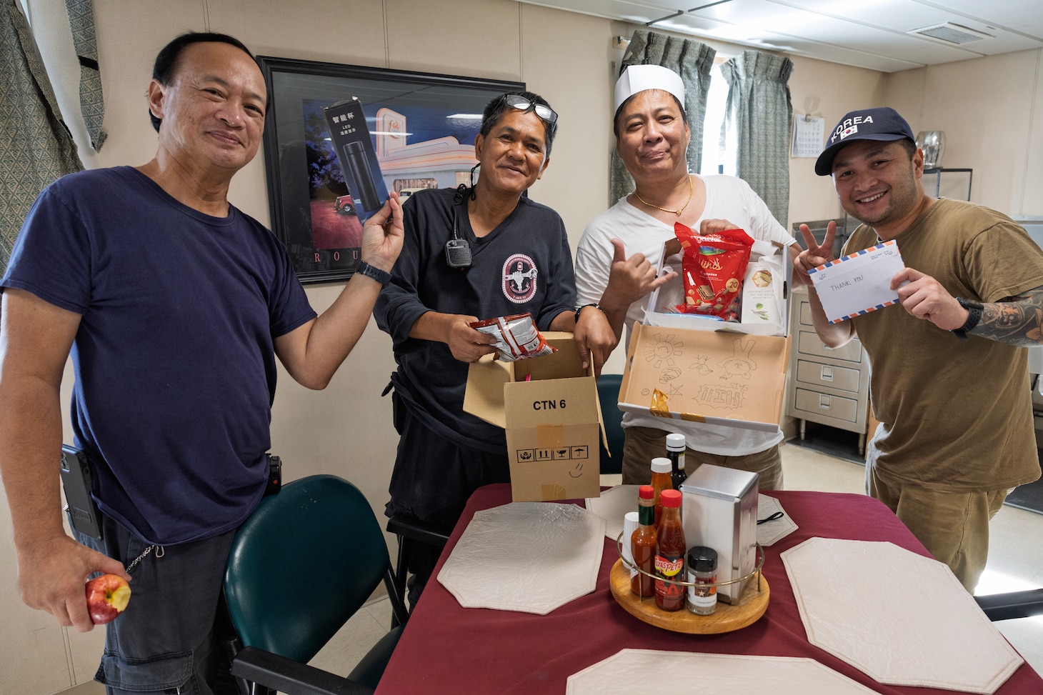 Ric Caniones, Jr., left, chief electrician, Armando Lucero, 2nd electrician, Sotero Garcia, chief cook, and Mark Laraza, 2nd cook, all with dry cargo and ammunition ship USNS Charles Drew (T-AKE 10), display the contents of their care package that was provided by the American Association of Singapore and the Stamford American International School in Singapore while aboard Charles Drew at Sembawang Wharves, Singapore, Nov. 21, 2024. (U.S. Navy photo by Grady T. Fontana)