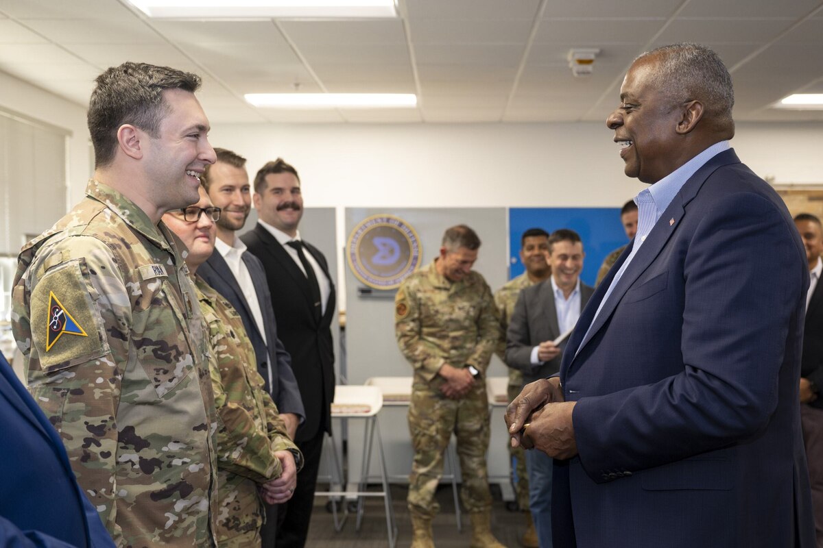 A man in business attire and a service member smiles at one another in a room. Service members and men in suits watch in the background.