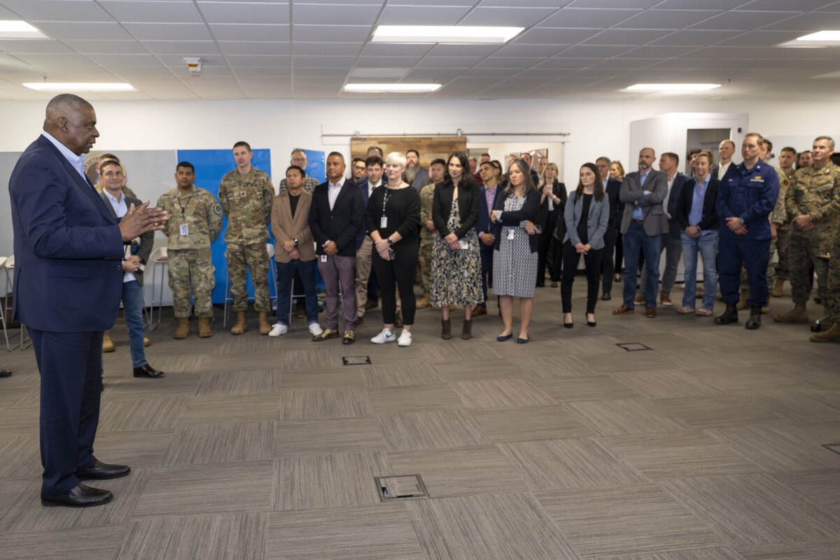 A man in business attire speaks to a group of service members in uniform and men and women in business attire inside a large room.