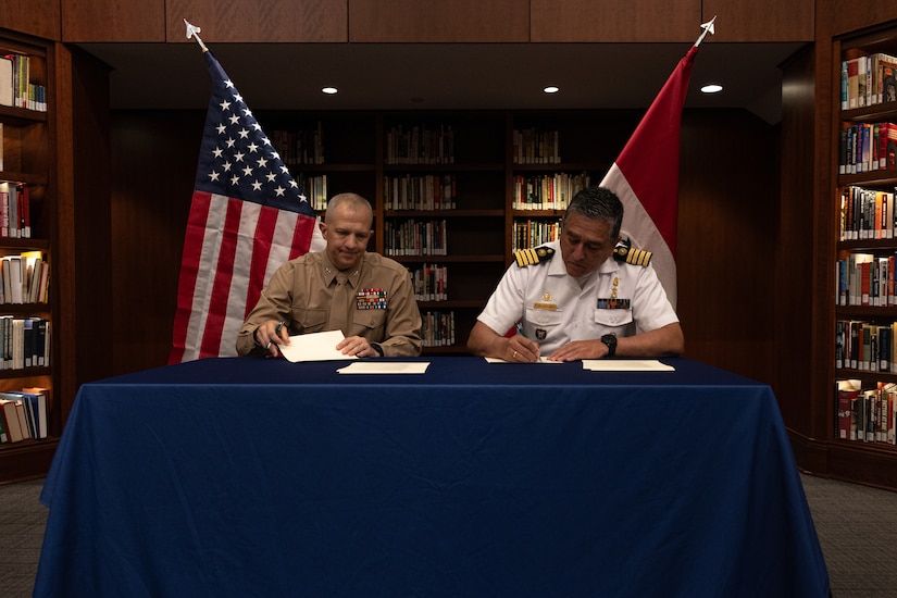 A Marine and a Peruvian officer sit next to each other at a table in front of their nations' flags and sign documents.