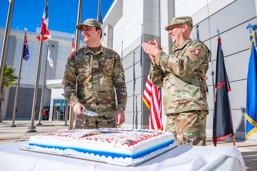 Two service members in military uniforms smile in front of a cake on a table outdoors. One holds a knife.