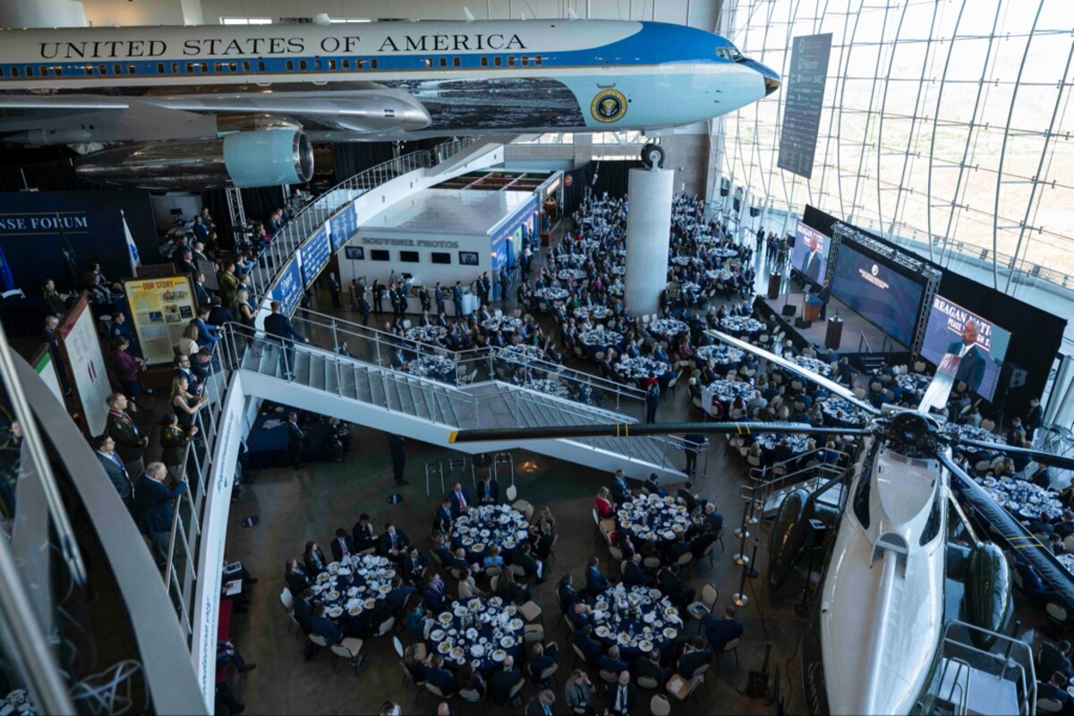 A man addresses a crowd of people seated at tables during an event. An aircraft model hovers above.
