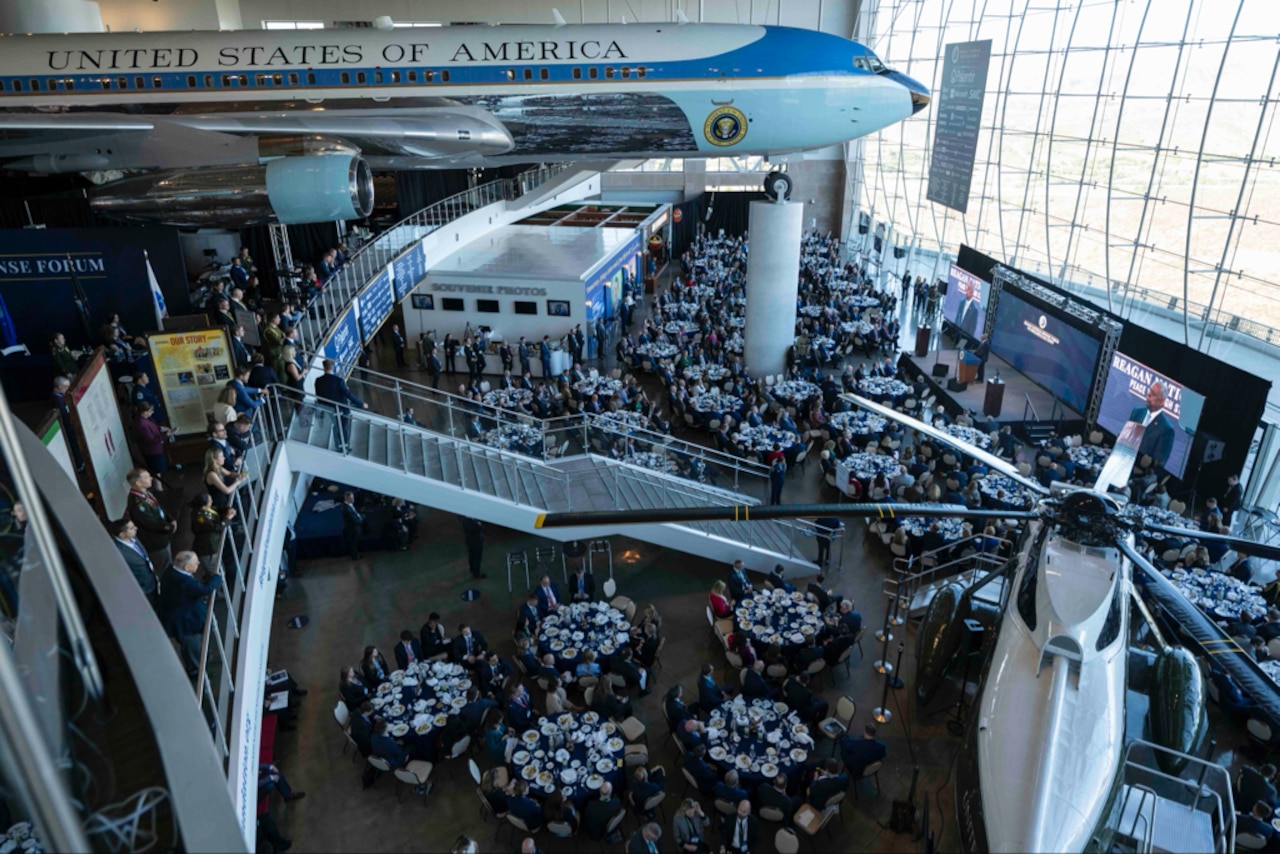 A man addresses a crowd of people seated at tables during an event. An aircraft model hovers above.