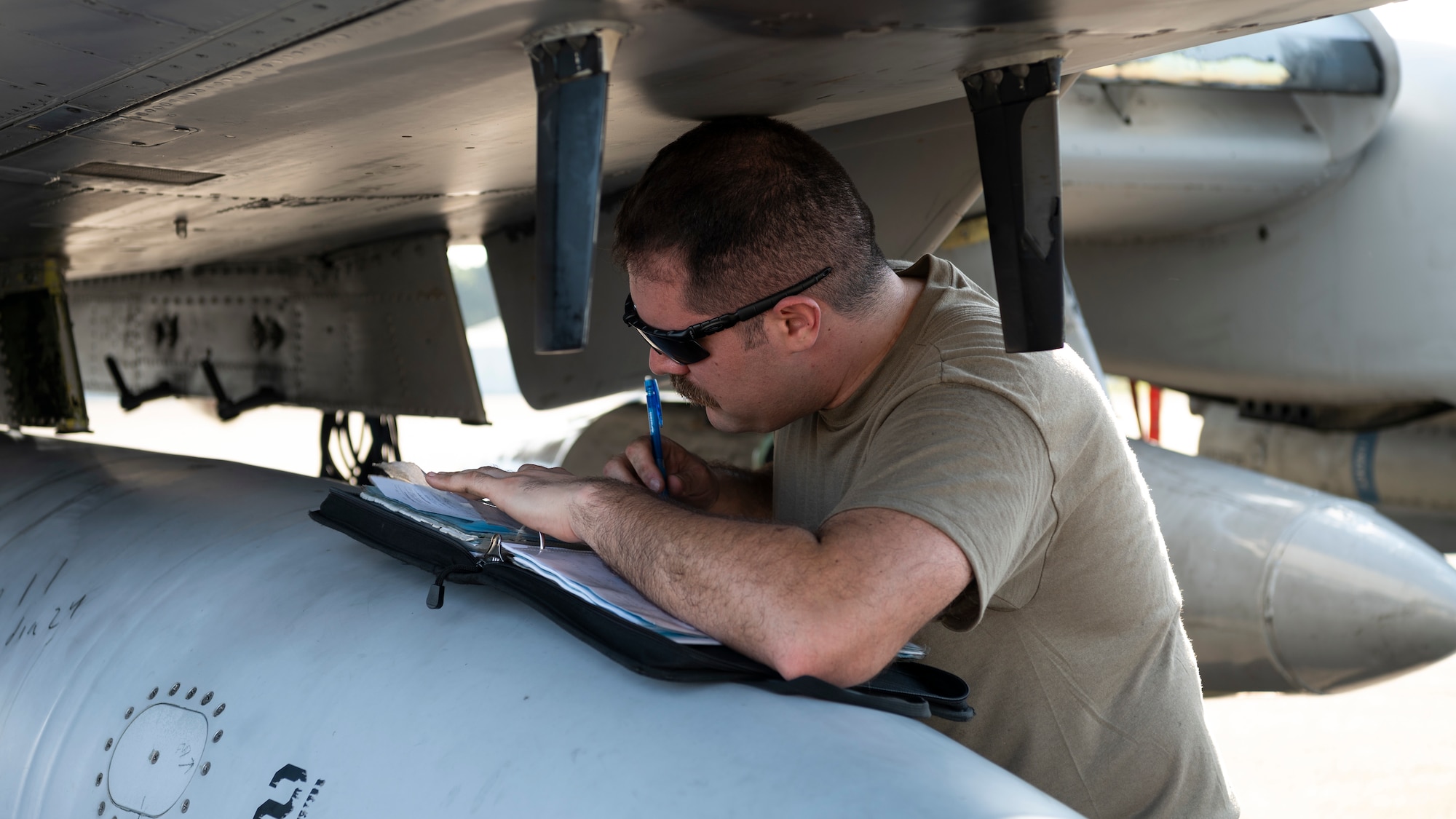 An Airman writes in a notebook underneath the wing of a fighter jet