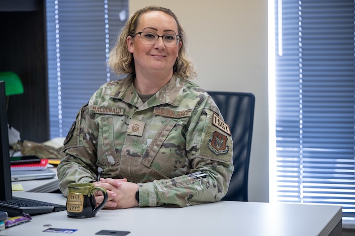 Master Sgt. Dava MacDonald sits at her desk, fingers intertwined and smiling.