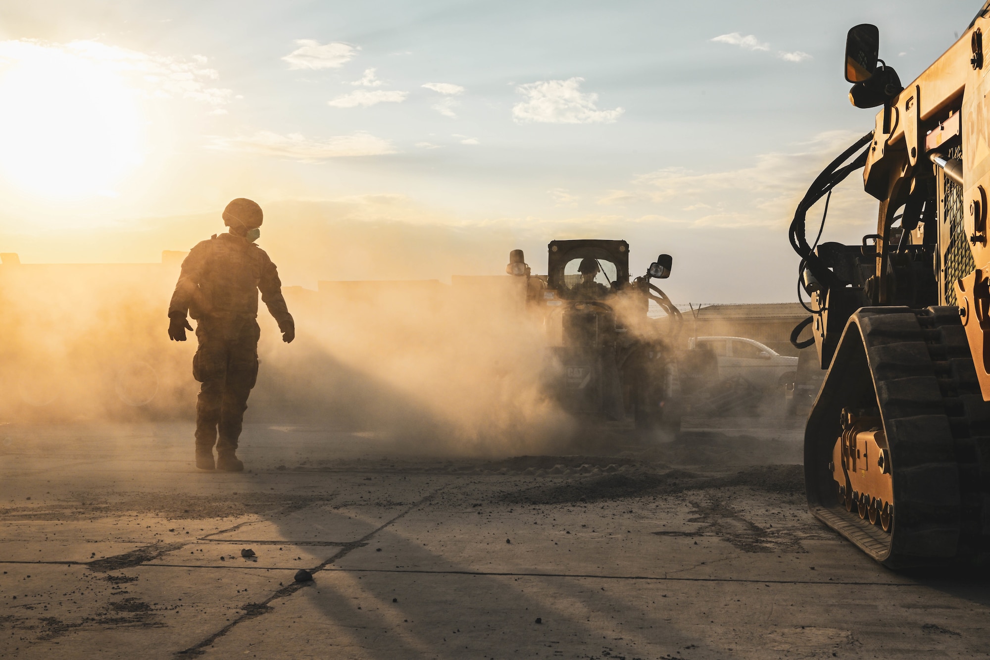 An Airman walks through a cloud of dust while other members operate heavy equipment.
