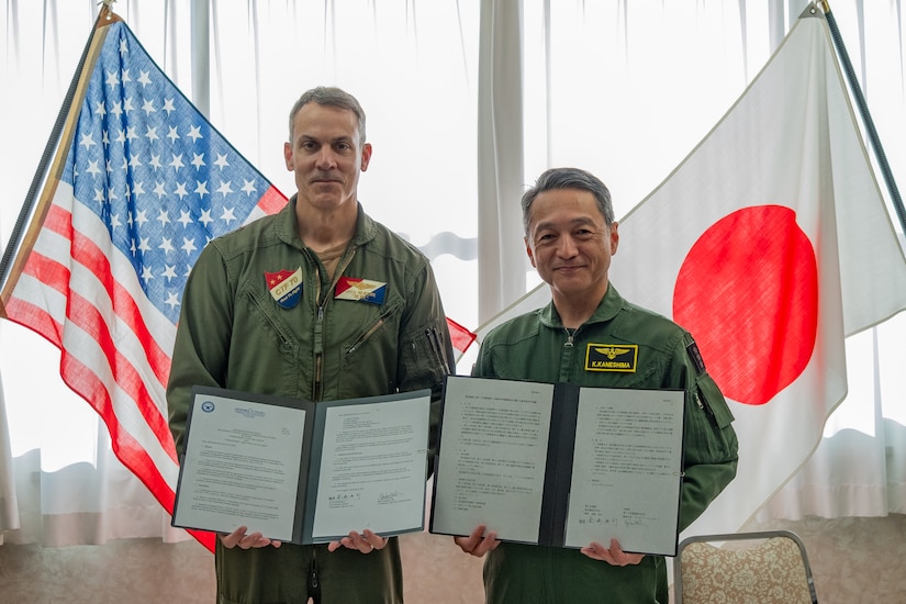 A U.S. and a Japanese navy officer stand holding signed documents in front of their nations' flags.