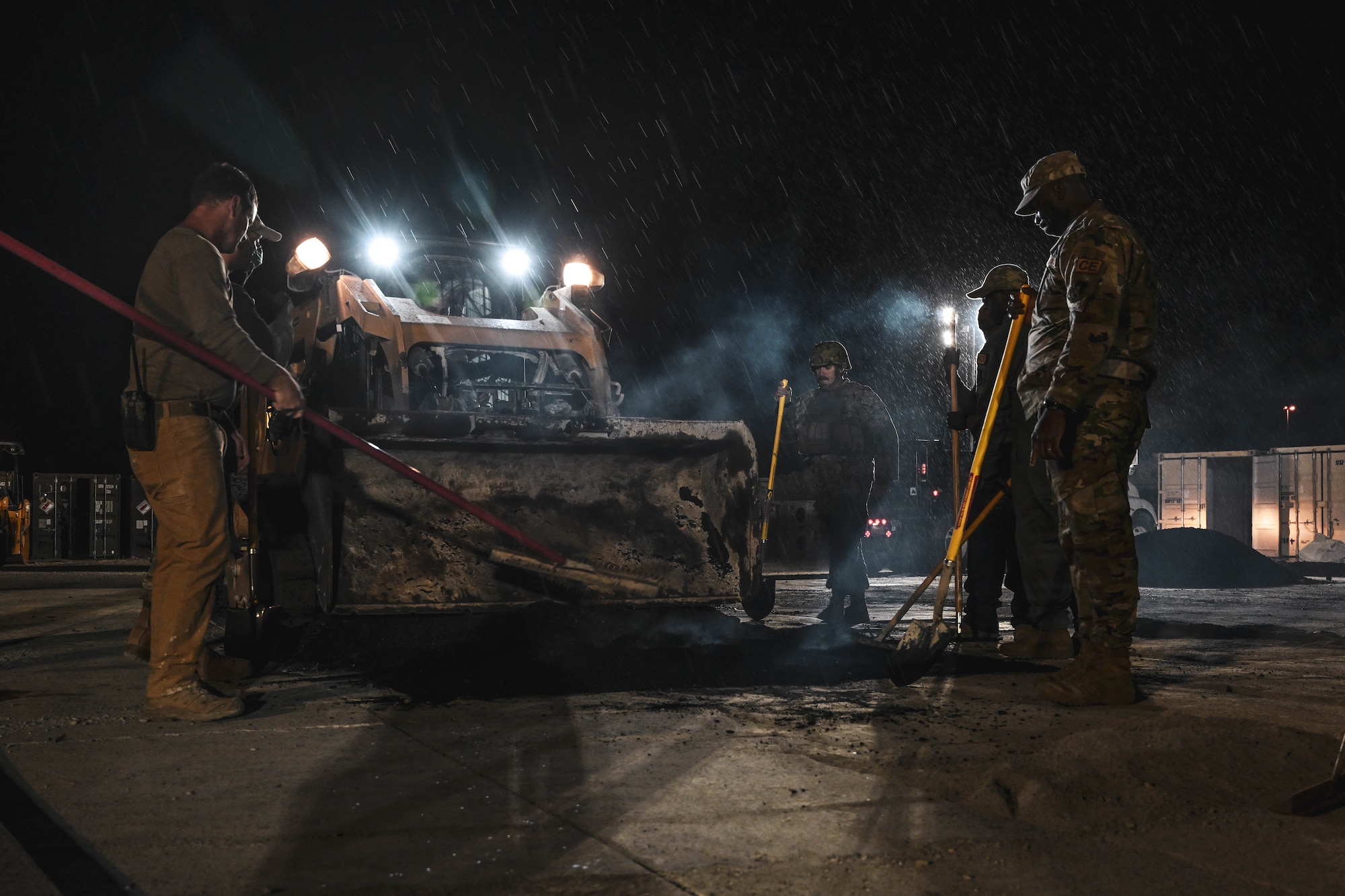 Military members look on as a heavy equipment operator pour asphalt.