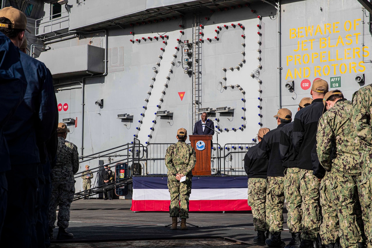 A civilian speaks to a large group of sailors on a ship.