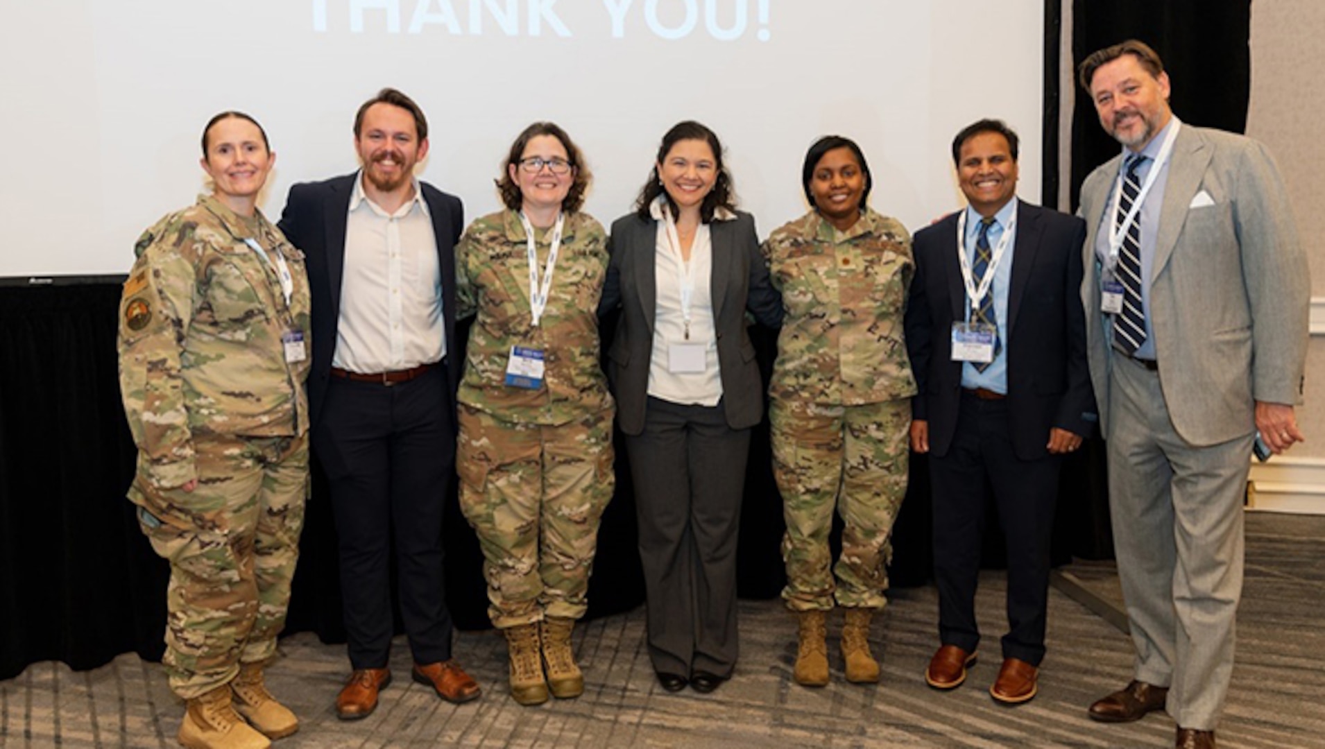 Three uniformed service members and four civilians in suits stand in front of projector screen.