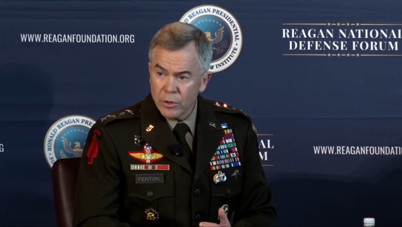 A man in a military uniform speaks from his seat on a stage.