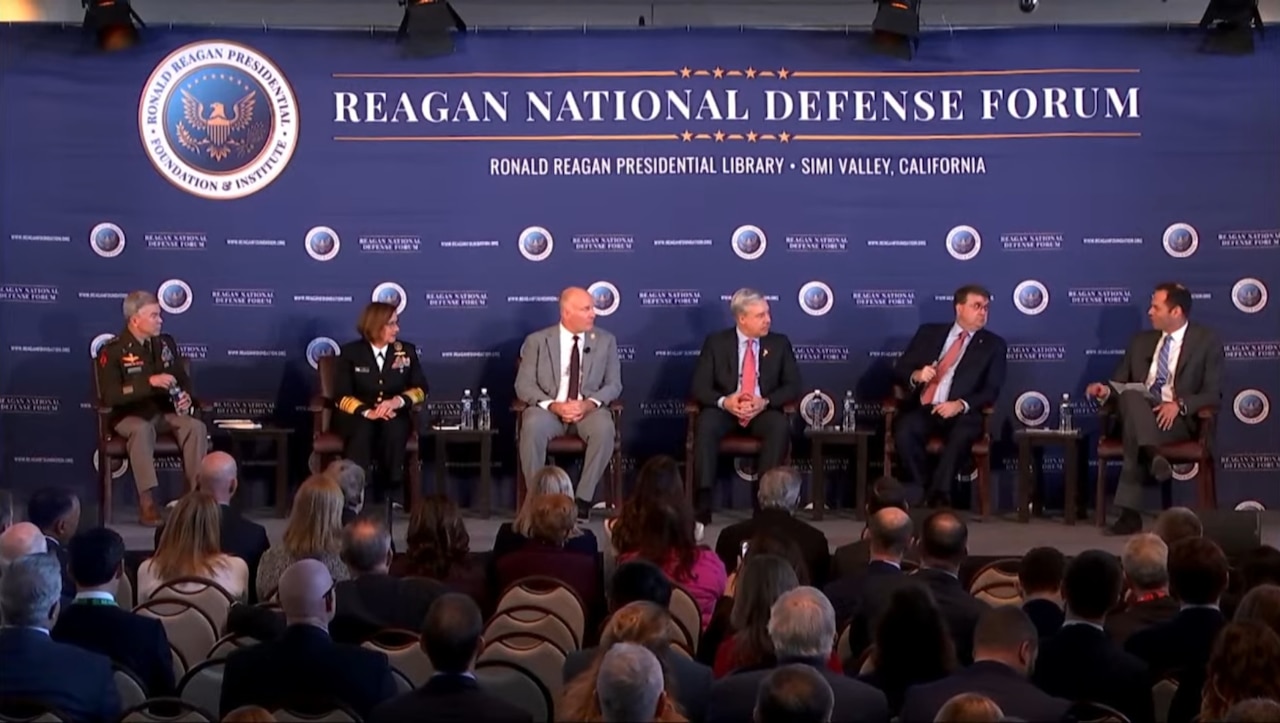 Six men and women hold a discussion from their chairs on stage while an audience listens in the foreground.
