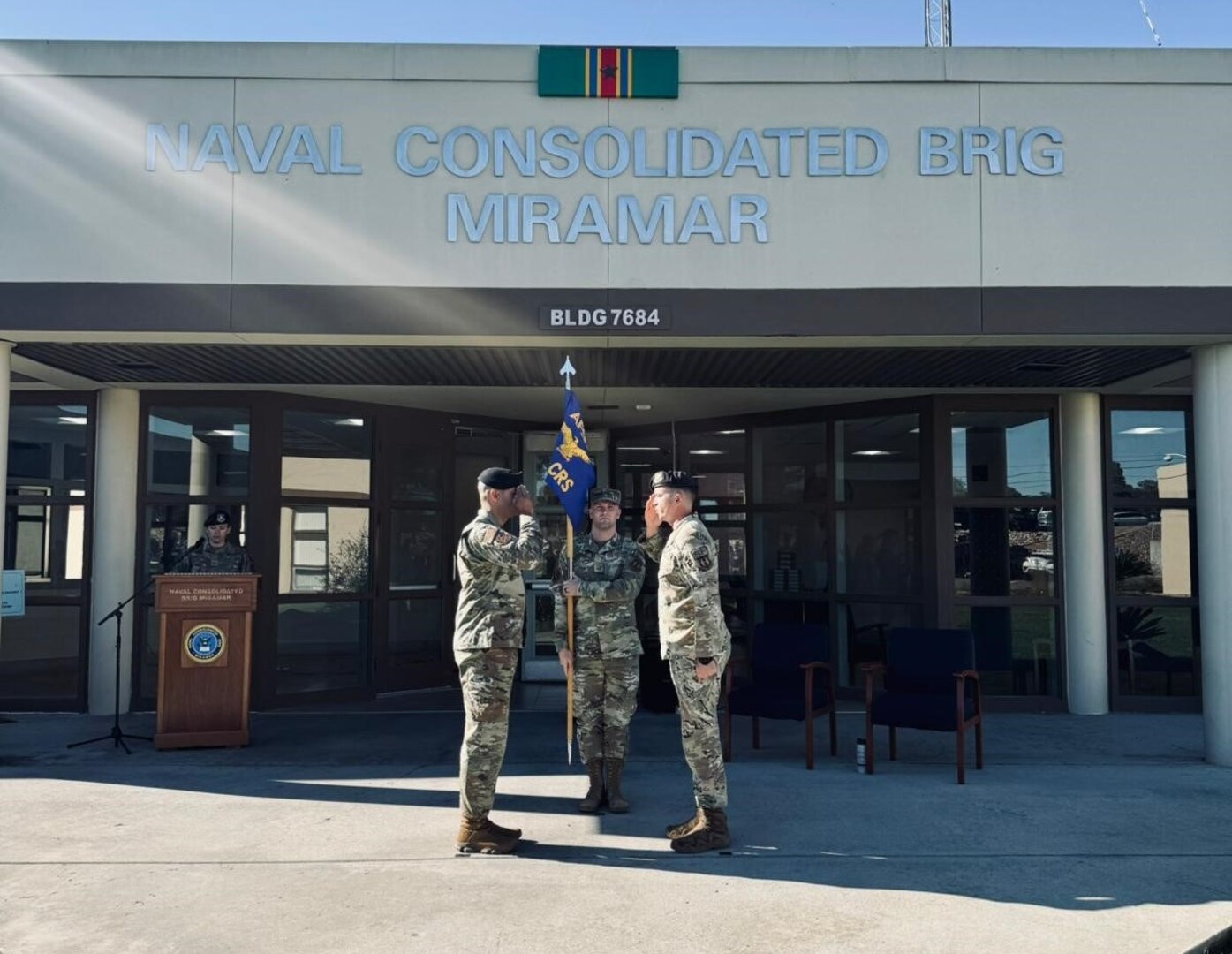 two officers salute each other outside of the corrections facility at Miramar while the first sergeant holds a guidon between them.