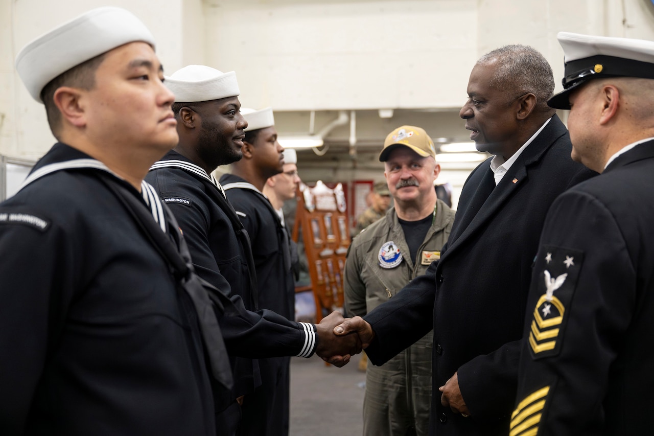 Secretary of Defense Lloyd J. Austin III shakes hands with a sailor standing with others aboard a ship.