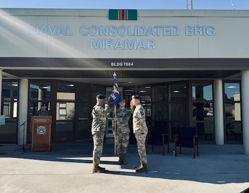 two officers salute each other outside of the corrections facility at Miramar while the first sergeant holds a guidon between them.