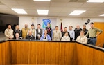 17 adult students stand behind a desk