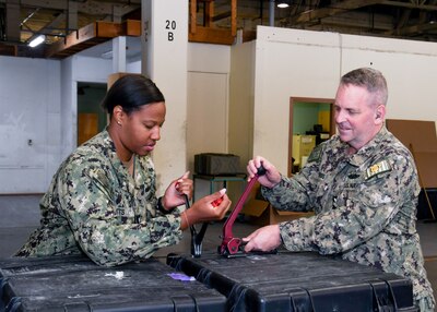 Two sailors in camo uniforms attach a binding strap to a box.