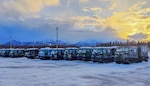 10 tracked vehicles with a forward cab and an attached passenger compartment sit on a snow covered parking lot with the sun coming up in the background. snow covered mountains can be seen in the distance.