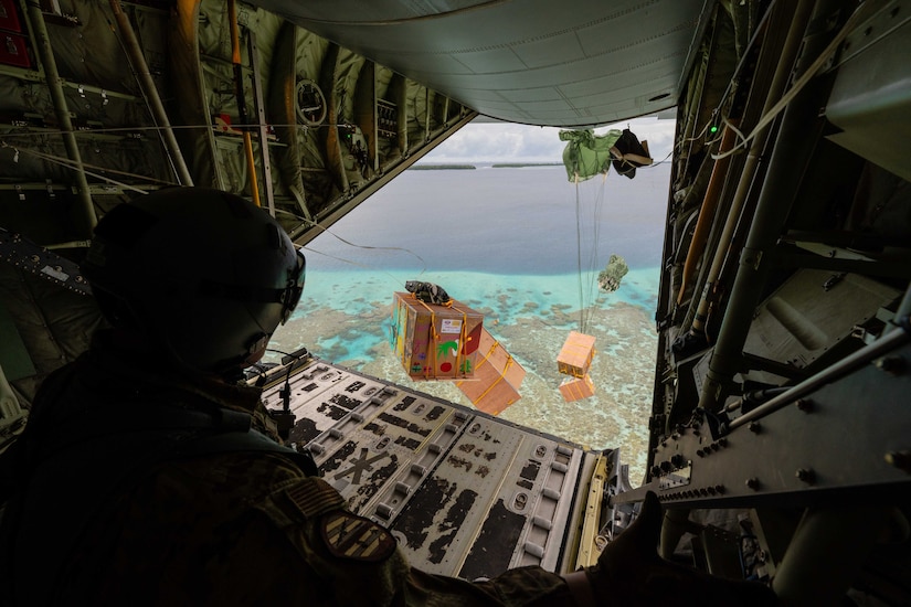 An airman watches four large boxes parachute out the back of a low-flying aircraft over water.