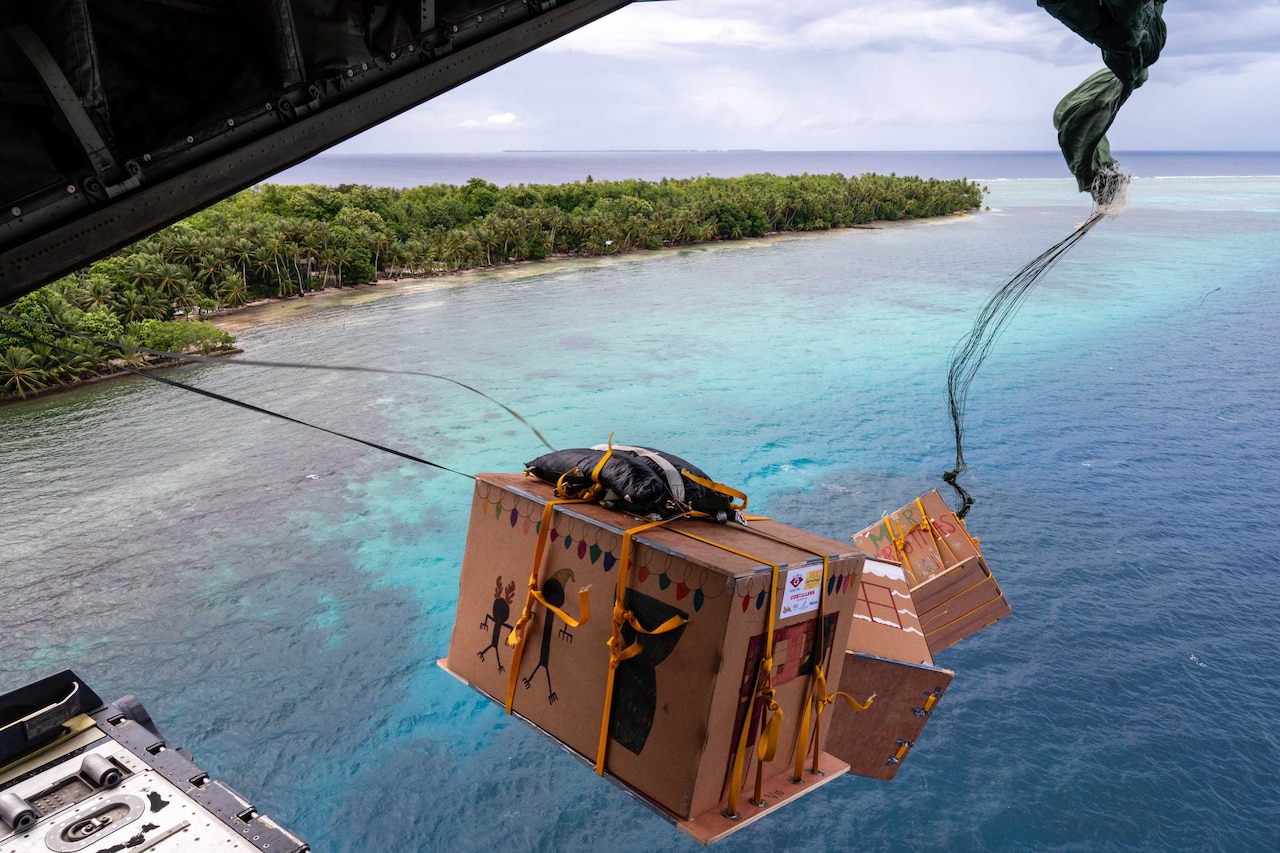 Three large boxes of supplies parachute out the back of a low flying aircraft over water and near land.