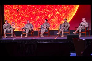 Five members in military uniform sit in chairs on a stage discussing topics.