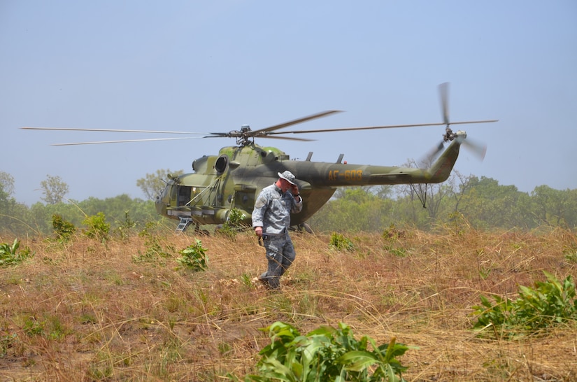 An Army civilian walks in a field with a helicopter in the background.