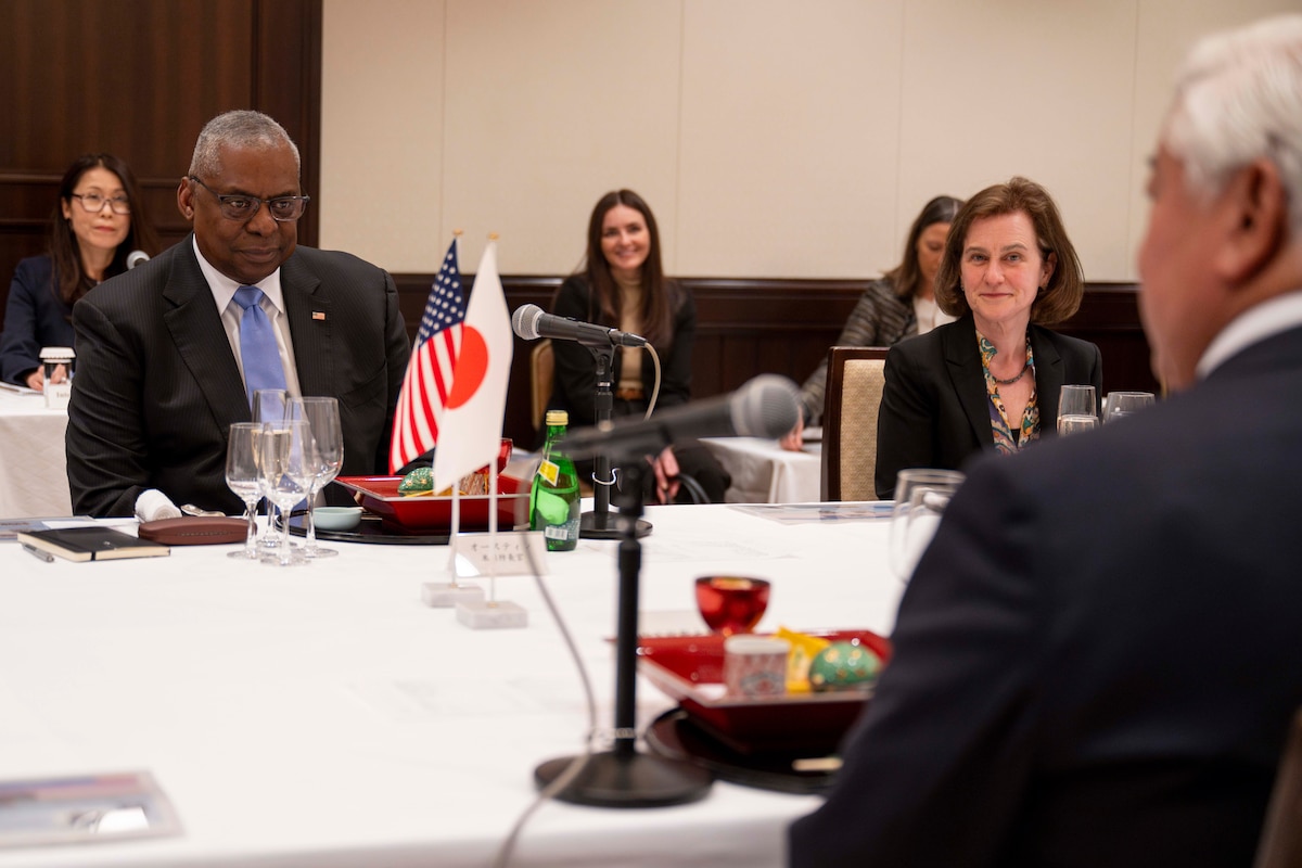 A man looks across a table at another man. A woman is seated beside him. Miniature U.S. and Japan flags are on the table.