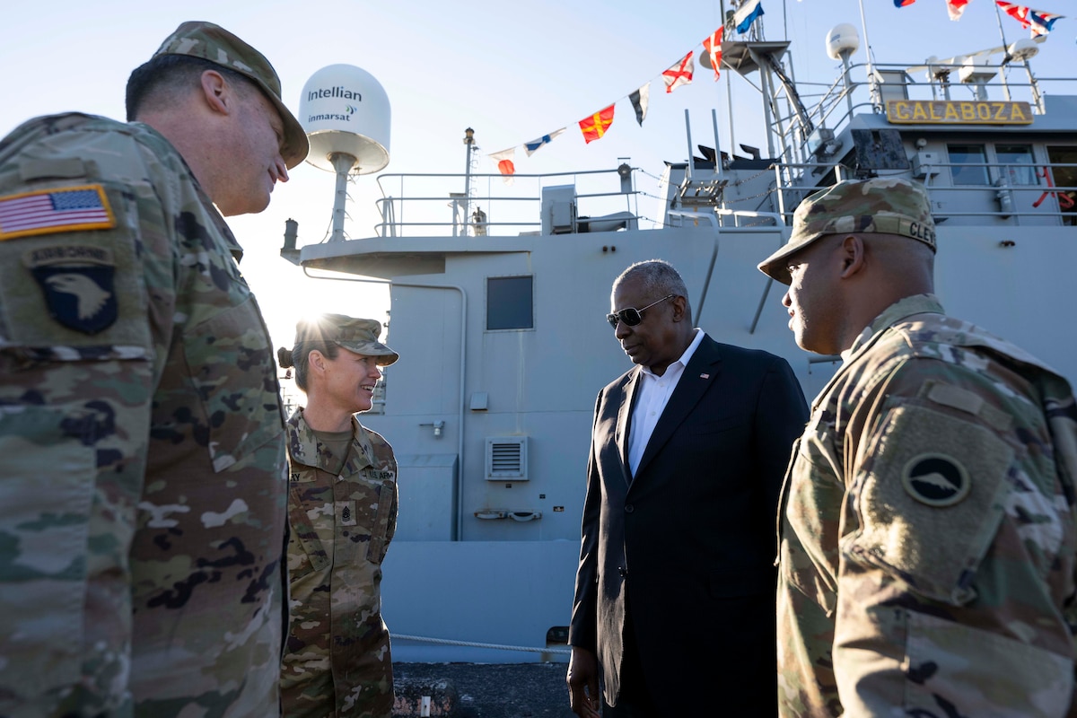 A group of soldiers greet a man in a suit. A military ship is in the background.