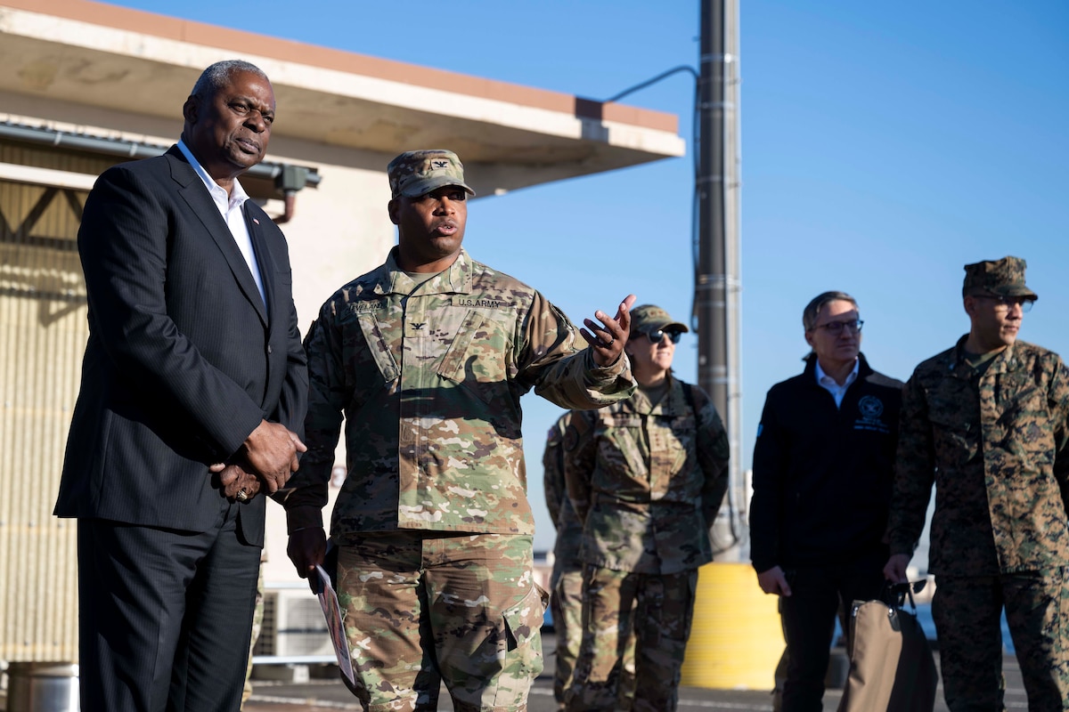 Two men, one in a military uniform, look off into the distance. Three soldiers and a man in civilian attire is standing behind them.