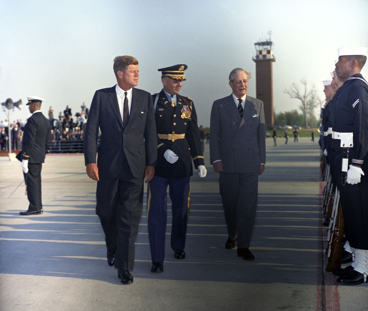 A man in uniform walks with two other men along an airport tarmac as several uniformed men stand at attention.