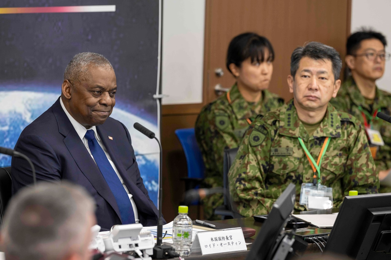 A seated man wearing a business suit smiles behind a microphone while a foreign service member looks on.
