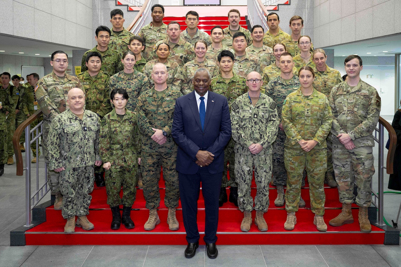 A man wearing a business suit stands in front of a large group of foreign and domestic service members for a photo.