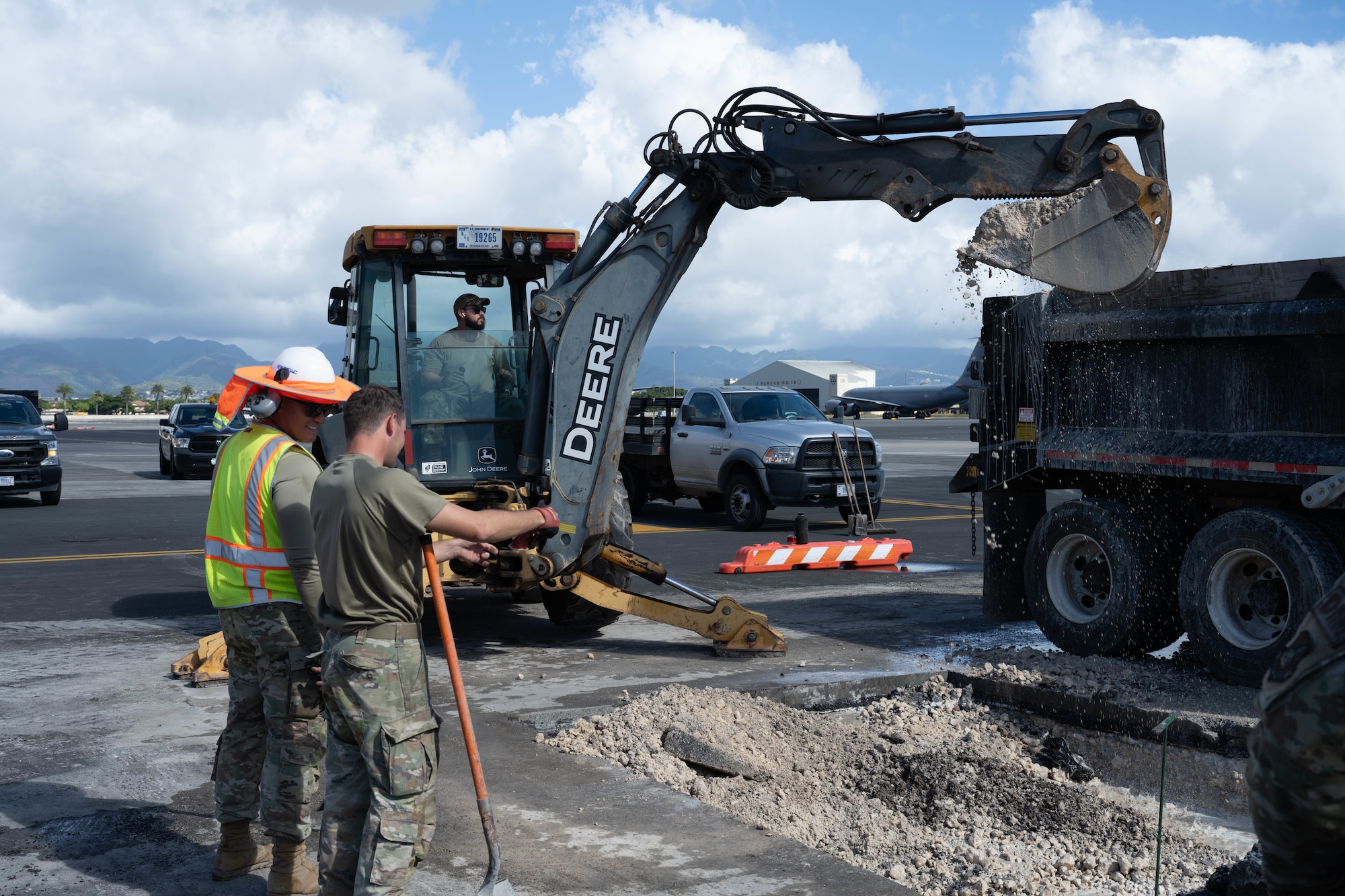 Airman operates an excavator and digs through wet concrete on the flightline surrounded by his team.