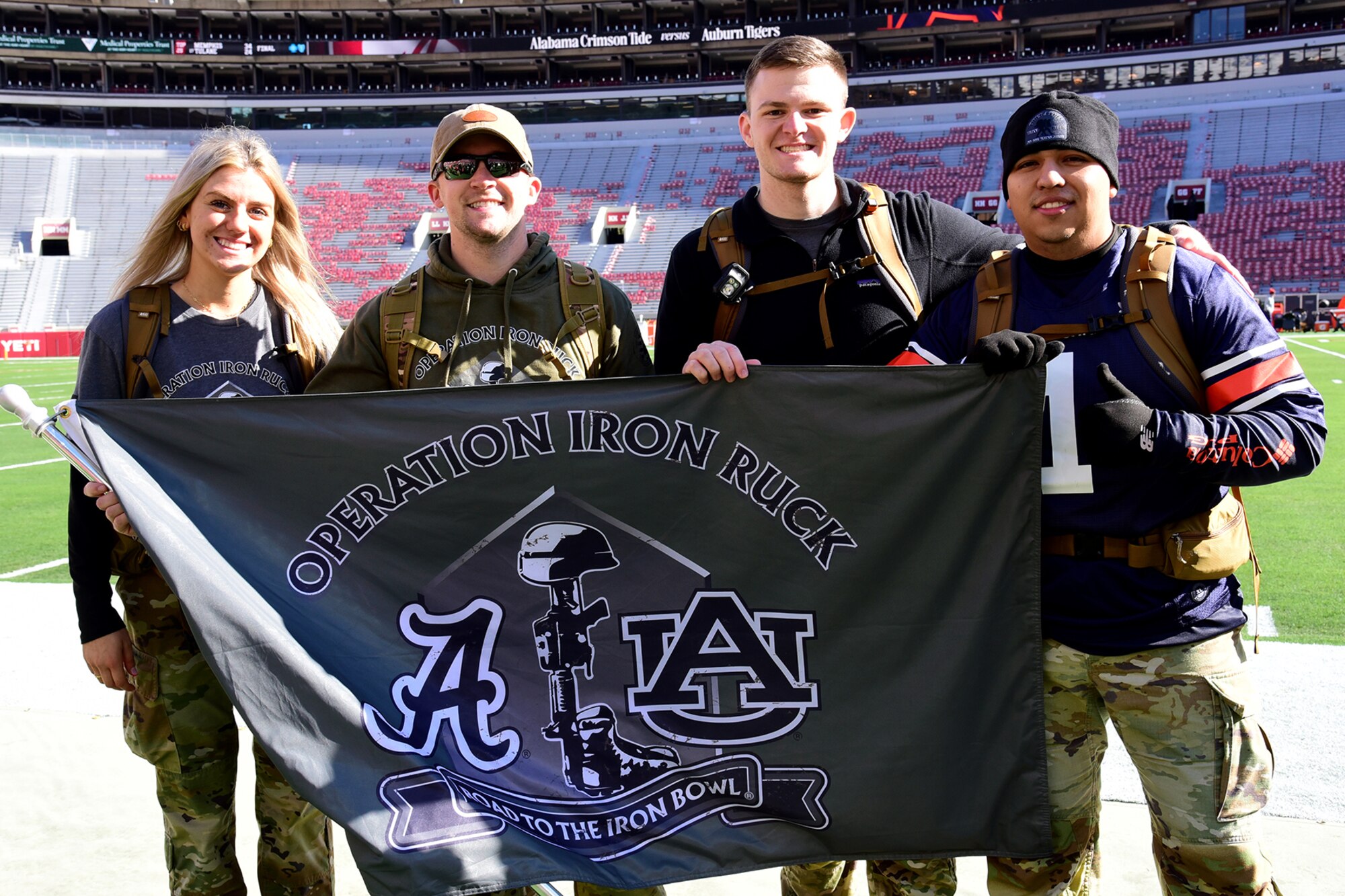 Four people stand together on a football field and hold a flag for Operation Iron Ruck.