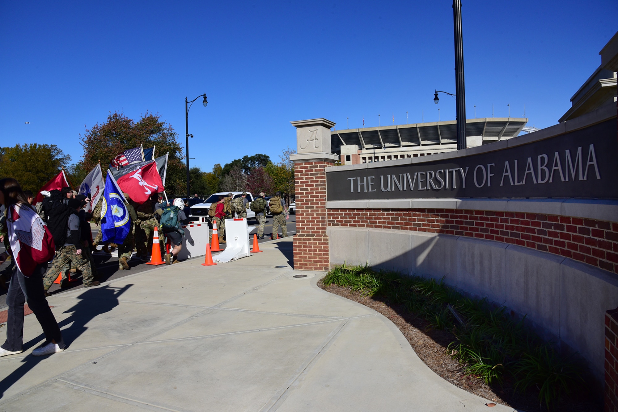 People walk together past the front sign for the University of Alabama, and some are carrying flags.