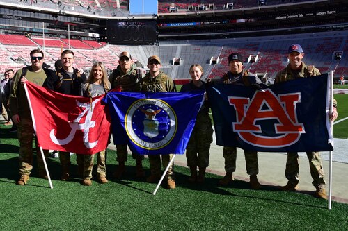 People stand in a group together holding flags on a football field. The flags, from left to right, are for Alabama University, the United States Air Force, and Auburn University.