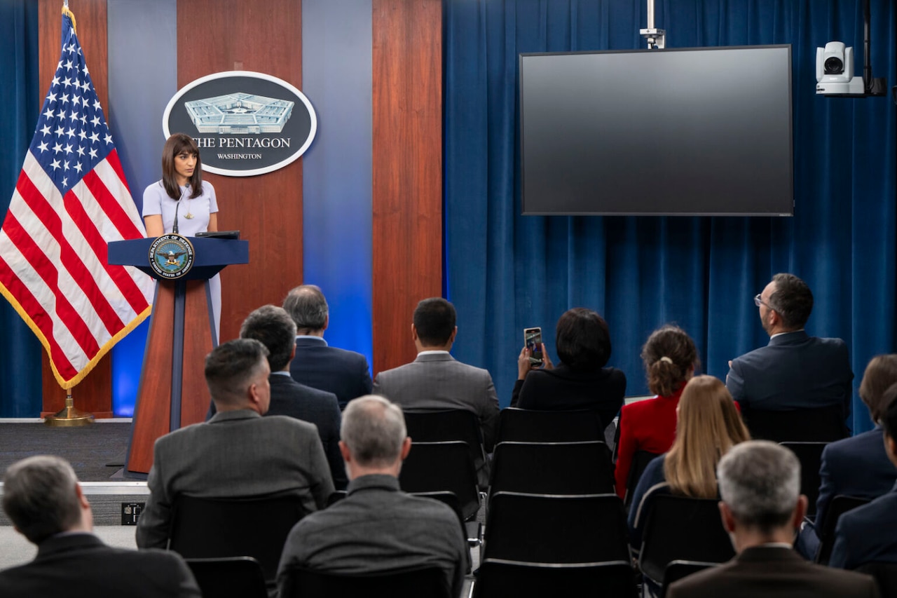 A woman is standing behind a lectern on a stage with the American flag and a wall-mounted logo showing a five-sided building that reads “The Pentagon” behind her. A group of people are sitting in chairs facing her, with their back to the camera.