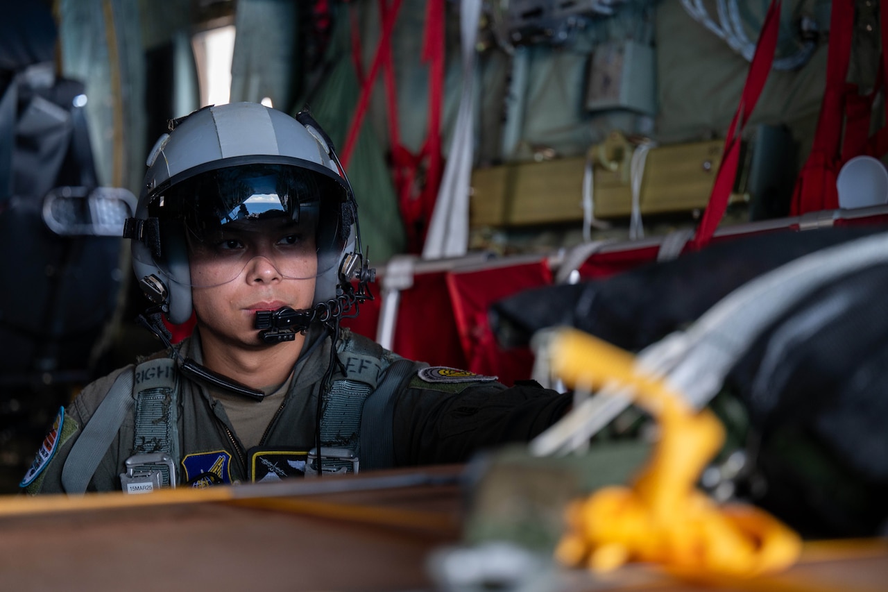 An airman with a helmet on sits inside the cargo bay of an aircraft.