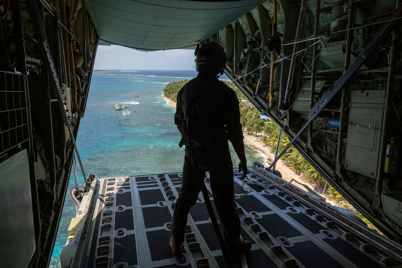 An airman watches out of the open back of an aircraft as supplies drop on parachutes over sea next to land.