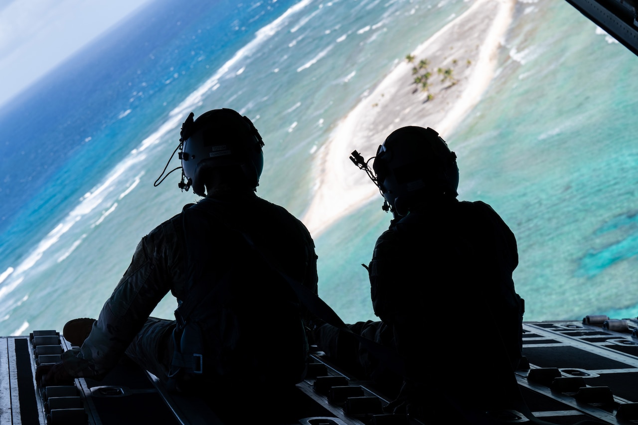 Two airman sit on the carbo bay of an aircraft and look out the back at the sea and an island.