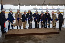 Ten people in hard hats are holding gold shovels standing behind a pit of sand