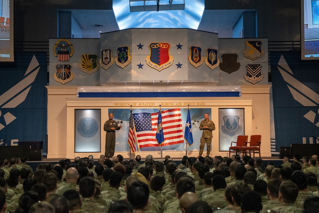 Photo shows two men in uniform on stage speaking to group of people in uniform.