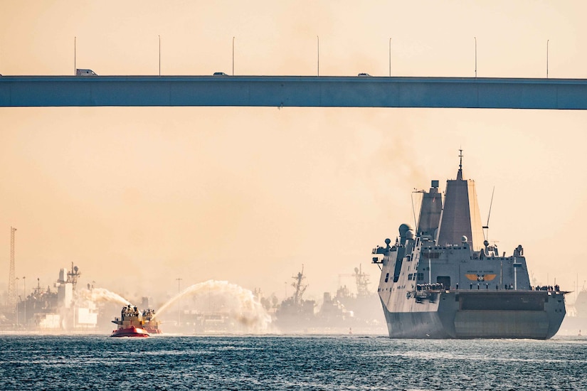 A military ship steams under a bridge.