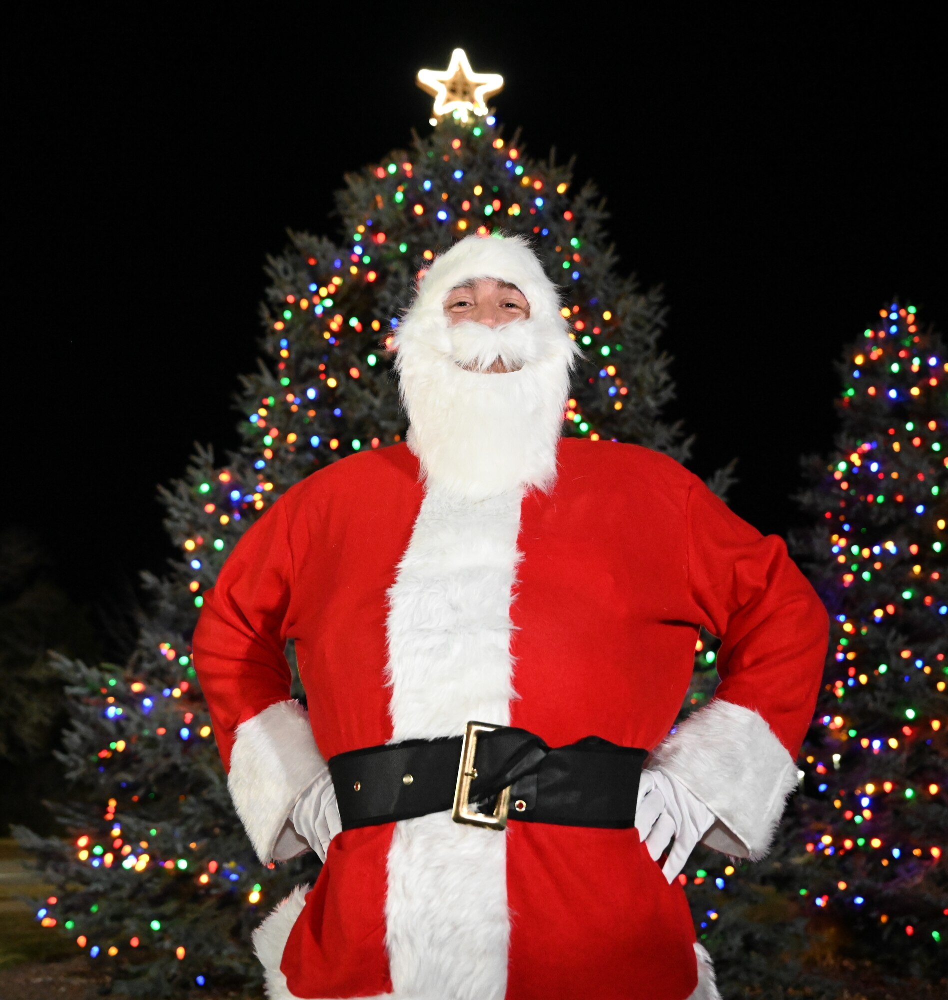 Santa Claus stands in front of two Christmas trees that have been decorated with lights and a topper.