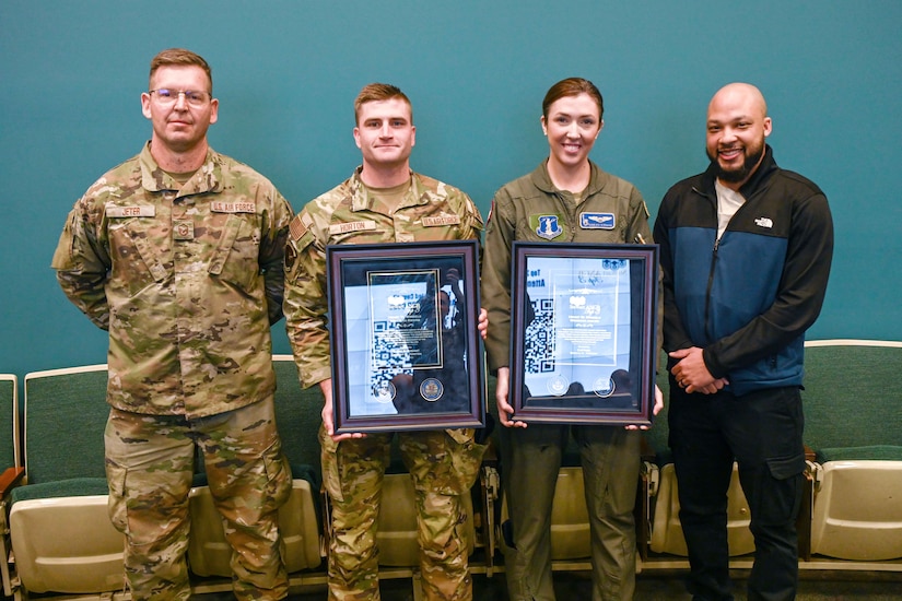 Four airmen stand next to each  while posing for a photo. The two in the center hold large awards.