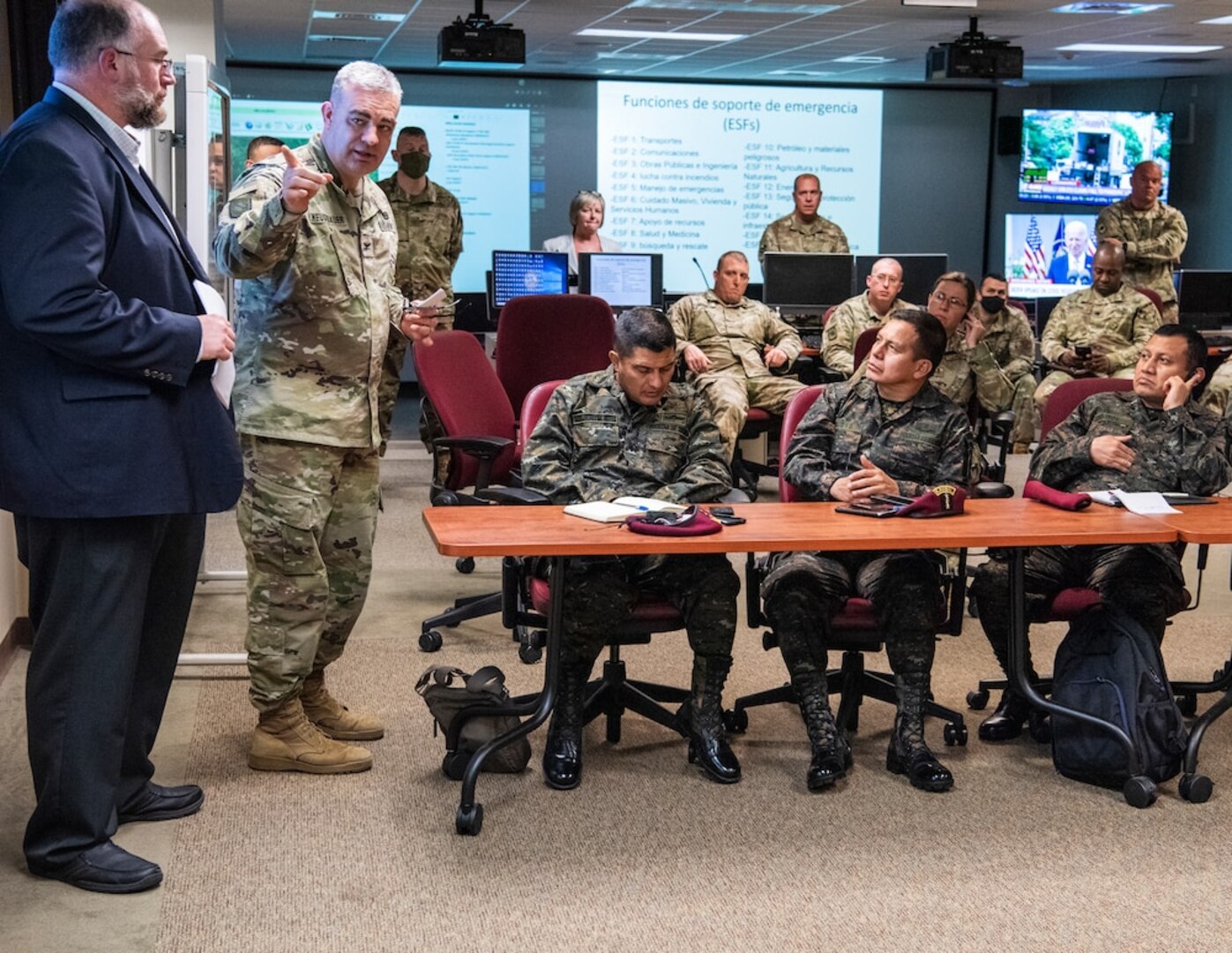 Soldiers sit in a classroom setting with screens in the background